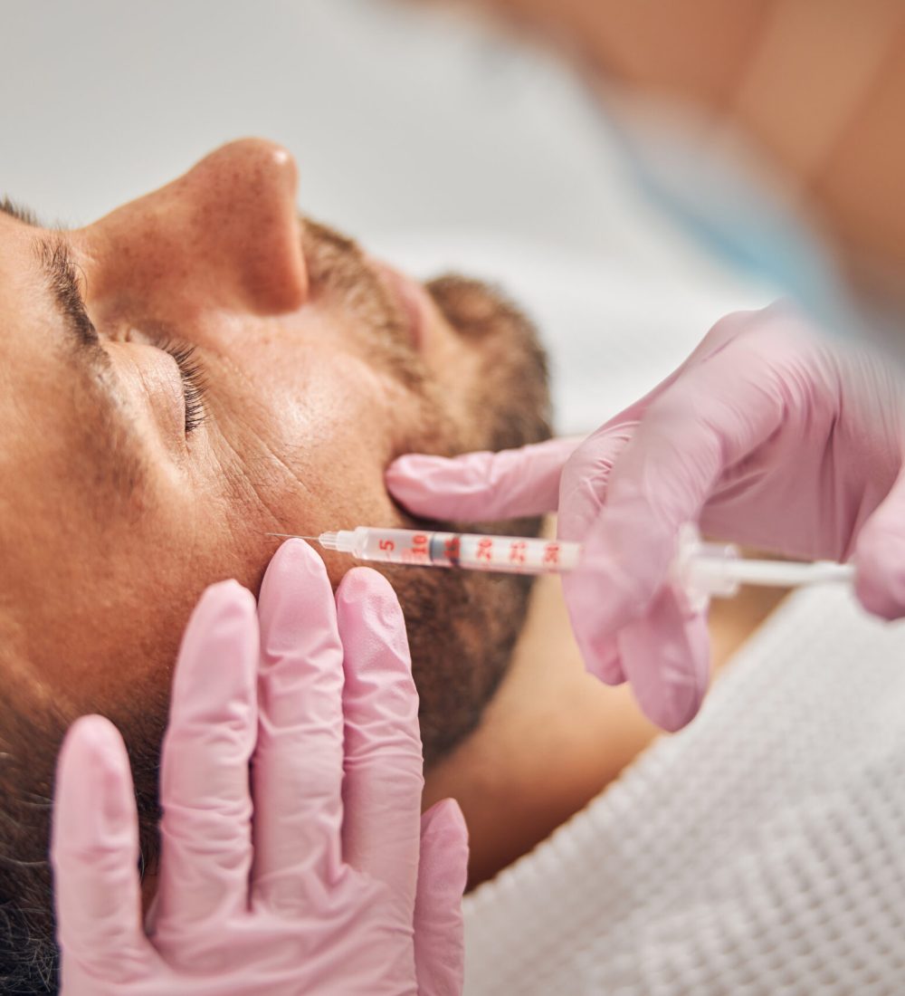 Close up of female cosmetologist in pink sterile gloves making injection in male forehead